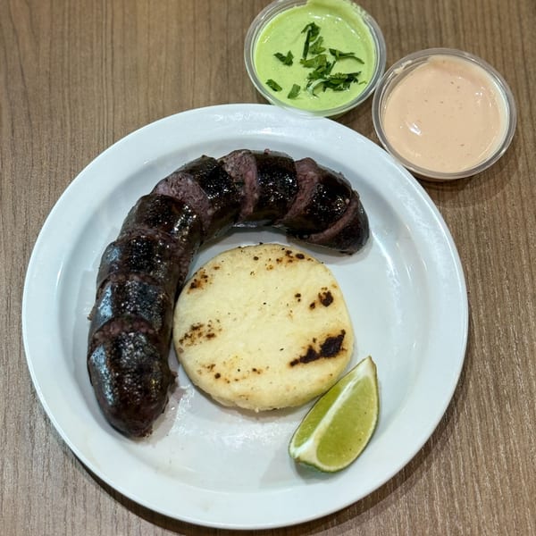 Nazca Express morcilla with an arepa, lime wedge, and two dipping sauces on a white plate.