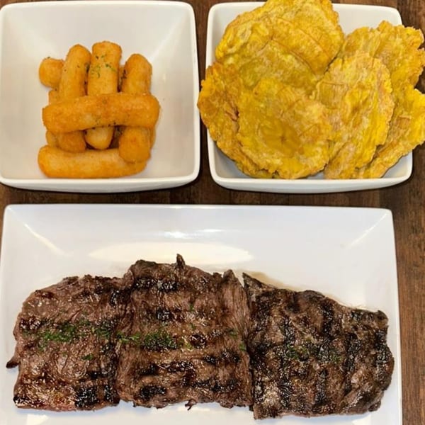 Grilled steak with fried yuca sticks and tostones on white plates from Nazca Express.