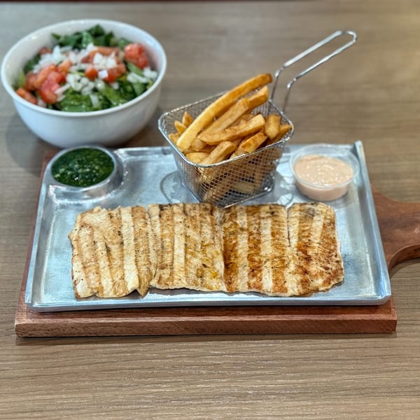 Grilled chicken, fries in a metal basket, green sauce, and a side salad on a wooden table.